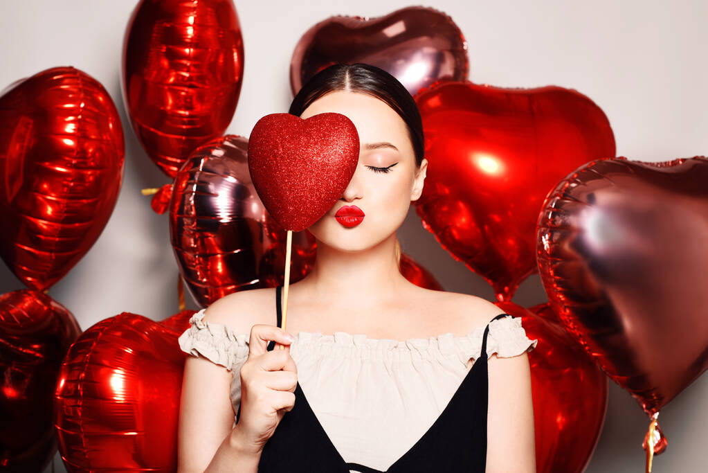 Beautiful girl, model on the background of balloons in the shape of a heart. Woman with red lips makeup, air kiss and good mood, love, feelings. Valentine's Day and March 8