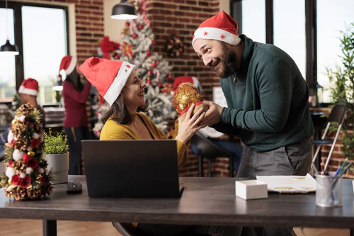 兴奋的女员工一边笑一边从戴着圣塔帽的同事那里收到圣诞礼物袋。快乐的高加索男人用圣诞节来问候同事，并在共同工作的空间里赠送礼物图片