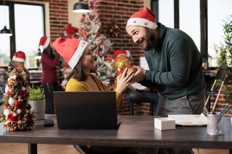 兴奋的女员工一边笑一边从戴着圣塔帽的同事那里收到圣诞礼物袋。快乐的高加索男人用圣诞节来问候同事，并在共同工作的空间里赠送礼物