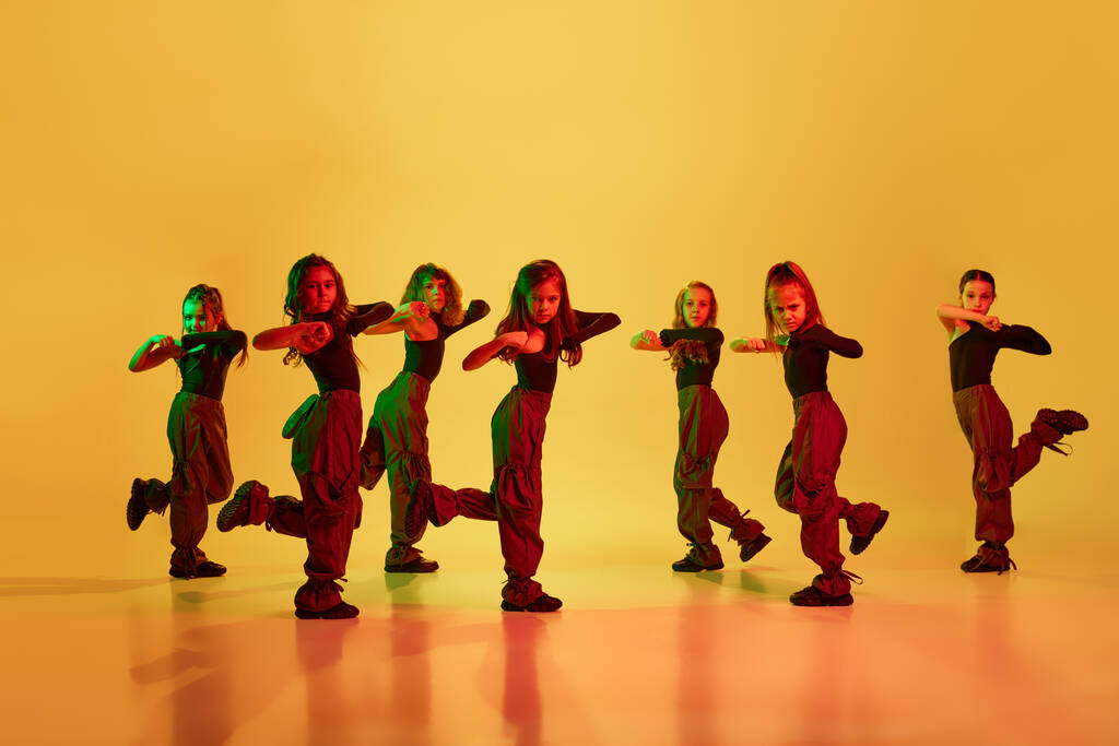 时髦的。一群孩子，穿着运动型休闲装的小女孩跳嘻哈舞，在霓虹灯下凝视着黄色的工作室背景跳舞。童年、业余爱好、体育生活方式的概念图片